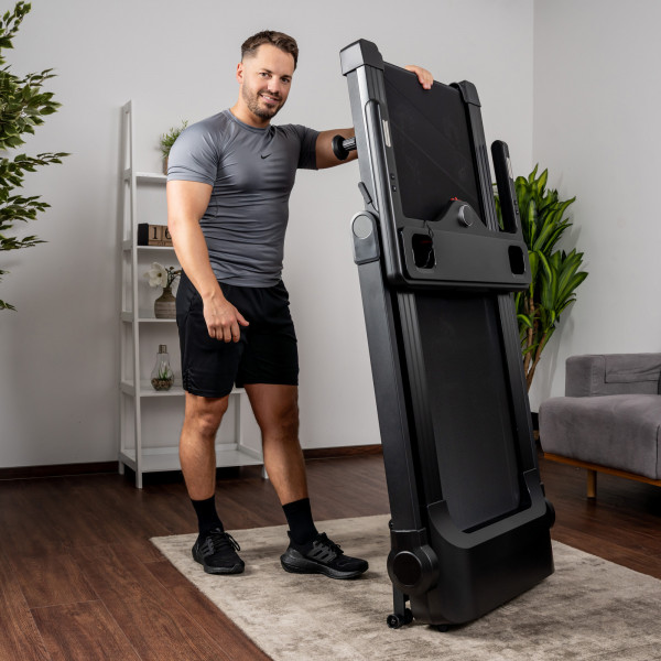 Eine Person in sportlicher Kleidung steht links neben dem Kettler Laufband Track 200, welches sie gerade aufgestellt hat. Mit der linken Hand greift die Person an das obere Ende des Laufbands.